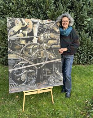 Fotografin und Grafikerin Beate Jeske steht neben einer Staffelei im Garten. Ihr Bild auf der Staffelei zeigt mehrere Zahnräder und Übersetzungsriemen.