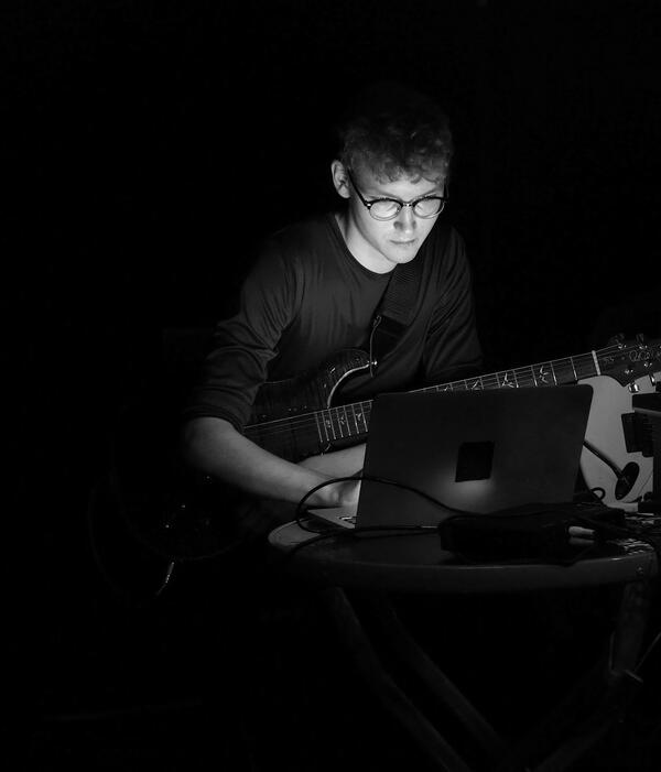Ein schwarz-weiß Foto. Ein Junge sitzt mit E-Gitarre im Arm am Laptop.