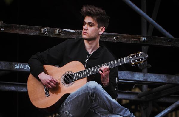 Ein junger Mann sitzt auf einem Gerüst. Er spielt auf seiner Gitarre.