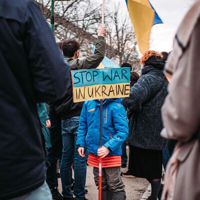 Menschen bei einer Demonstration. In der Mitte steht ein Kind mit einem Schild, auf dem "Stop den Krieg in der Ukraine" auf englisch geschrieben steht.