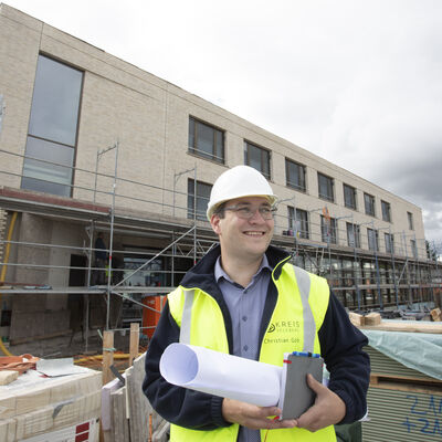 Ein Kreismitarbeiter mit gelber Weste guckt sich auf einer Baustelle um, trägt einen weißen Helm und hat Pläne bei sich.