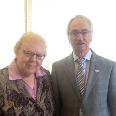 Ein Bild von einer Frau und einem Mann. Die Frau trägt ein mit Blumen besticktes Oberteil und darunter eine pinke Bluse. Der Mann trägt einen grauen Anzug mit einer blau gestreiften Krawatte.