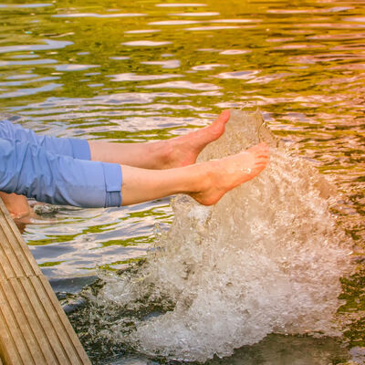 Eine Person sitzt auf einem Steg an einem See und spritzt mit den Füßen im Wasser.
