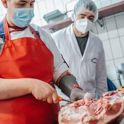 Ein Mann schneidet ein Stück Fleisch und wird dabei von einem Kreismitarbeiter beobachtet.