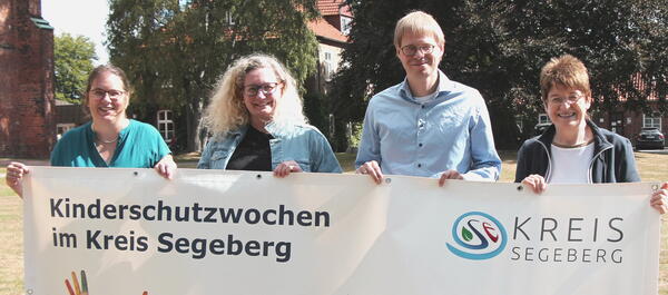 Vier Personen halten einen weißen Banner mit der Aufschrift "Kinderschutzwochen im Kreis Segeberg". Im Hintergrund sieht man Bäume und Gebäude.