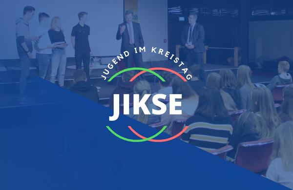 Das Logo JIKSE vor einem blauen Hintergrund. Im Hintergrund sind auch ganz schwach der Landrat, der Kreispräsident und viele Schüler*innen zu sehen.