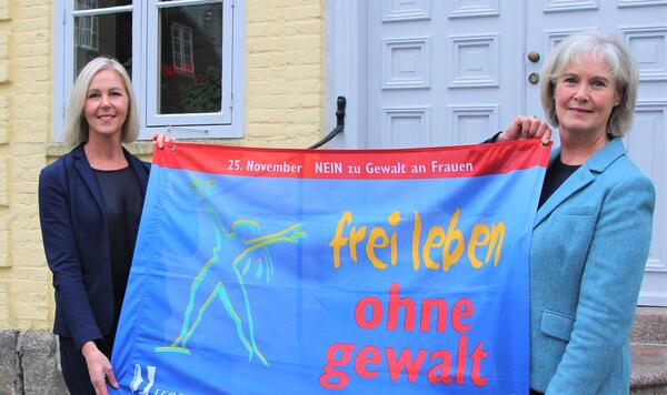 Zwei Frauen stehen vor einem gelben Haus und halten ein blaues Plakat hoch. Auf dem Plakat steht "Frei leben ohne Gewalt".