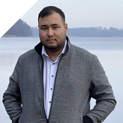 Ein l&auml;chelnder Mann steht vor einem See und tr&auml;gt eine graue Jacke.