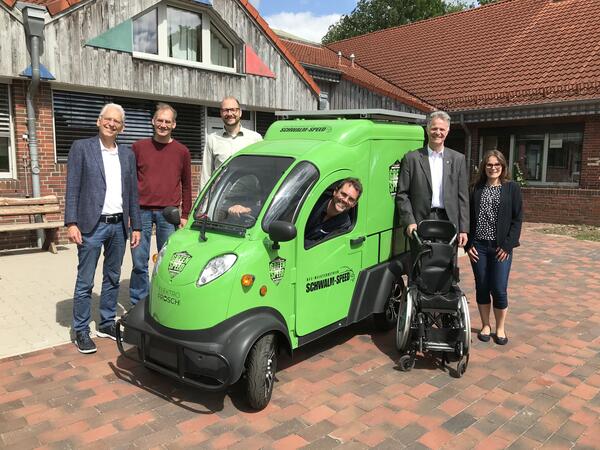 Fünf Menschen stehen neben einem grünen Elektro-Auto auf dem Vorplatz des Förderzentrums. Ein Mann sitzt in dem Auto und guckt zum Fenster hinaus.