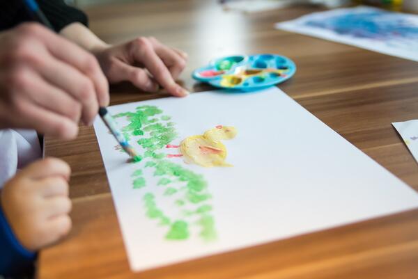 Eine Erwachsenenhand und Kinderhände tuschen ein Bild von einem Küken auf einer grünen Wiese.