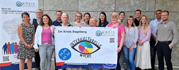 Einige Personen haben sich vor einer Hauswand versammelt und halten ein Plakat hoch. Auf dem Plakat zu sehen ist ein Auge mit einer bunten Iris und der Aufschrift "Interkulturelle Woche" 24.09.-01.10.