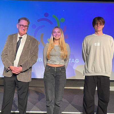 Ein Mann im Anzug steht mit einer Schülerin und einem schüler auf einer Bühne. Im Hintergrund ist das Logo von Jugend im Kreistag auf einer Leinwand.