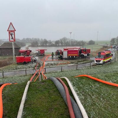 Verschiedene Schläuche liegen auf einer Wiese und führen über einen Deich. In dessen Hinterland stehen Einsatzwagen der Feuerwehr vor einer überfluteten Wiese.