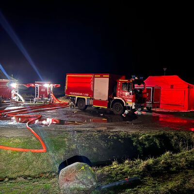 Verschiedene Schläuche liegen auf einer Wiese und führen in Richtung Deich. In dessen Hinterland stehen Einsatzwagen der Feuerwehr. Überall sind große Pfützen zu sehen.