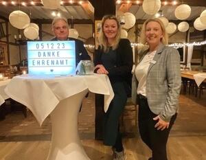 Zwei Frauen und ein Mann stehen an einem Stehtisch in einem Restaurant. Auf dem Stehtisch steht ein Schild mit einer Danksagung an ehrenamtlich Tätige.