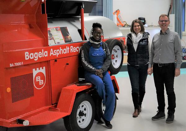 Zwei Frauen und ein Mann stehen in einer Halle vor einem roten Auto und lächeln.