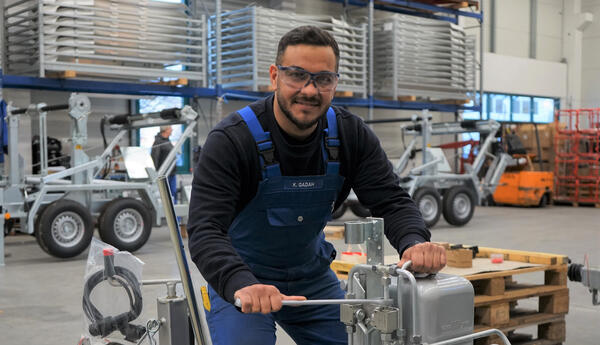 Ein Mann in einer blauen Latzhose arbeitet in einer Halle an einer kleineren Maschine mit Hebel.