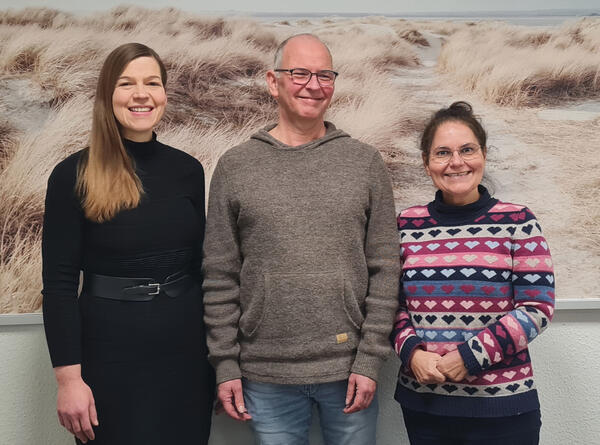 Zwei Frauen und ein Mann stehen vor einer Wand. An der Wand hängt ein großes Bild mit einer Dünenlandschaft und Meer.