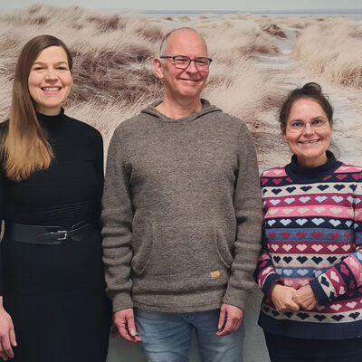 Zwei Frauen und ein Mann stehen vor einer Wand. An der Wand hängt ein großes Bild mit einer Dünenlandschaft und Meer.