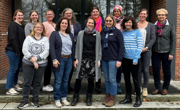 13 Frauen stehen vor einem Gebäudeeingang und lächeln.
