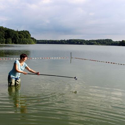 Eine Frau steht in einem See und entnimmt eine Wasserprobe. Dafür hält sie einen langen Stab, an dessen Ende das Probengefäß angebracht ist.