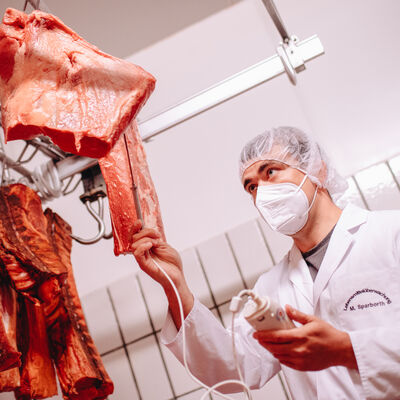 Eine Person mit Kittel, Haarnetz und Maske kontrolliert die Qualität vom Fleisch.