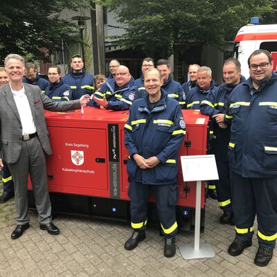 Viele Einsatzkräfte des Technischen Hilfswerks mit blauen Uniformen stehen mit Landrat Jan Peter Schröder neben einer Art roten Kiste.