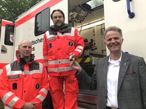 Zwei Männer in roter Einsatzkleidung stehen zusammen mit Landrat Jan Peter Schröder vor der Seitentür eines DRK-Einsatzwagens.