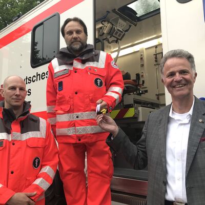 Zwei Männer in roter Einsatzkleidung stehen zusammen mit Landrat Jan Peter Schröder vor der Seitentür eines DRK-Einsatzwagens.