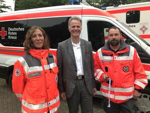 Eine Frau und ein Mann in roter Einsatzkleidung stehen gemeinsam mit Landrat Jan Peter Schröder vor einem Einsatzwagen des DRK.