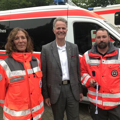 Eine Frau und ein Mann in roter Einsatzkleidung stehen gemeinsam mit Landrat Jan Peter Schröder vor einem Einsatzwagen des DRK.