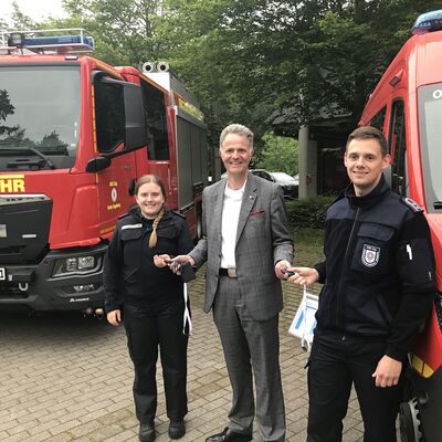Eine Frau und ein Mann in dunkelblauer Einsatzkleidung stehen gemeinsam mit Landrat Jan Peter Schröder vor zwei großen Einsatzwagen der Feuerwehr.