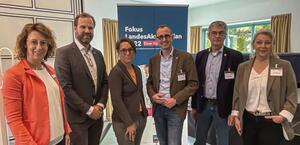 Drei Frauen und drei Männer stehen in einem Raum im Halbkreis und lachen in die Kamera.