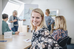Mehrere Frauen sitzen und stehen an einem Tisch. Im Vordergrund lächelt eine blonde Frau in die Kamera.