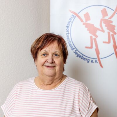 Eine ältere Dame mit kurzen Haaren steht vor einem Plakat mit tanzenden Figuren.