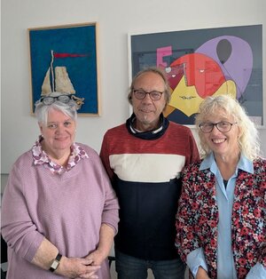 Die drei Beauftragten stehen vor mehreren gemalten Bildern und lächeln.