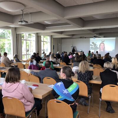 Mehr als zehn Menschen sitzen einem großen Raum in mehreren Stuhlreihen und sehen auf eine Beamer-Projektion.