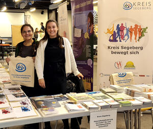 Zwei Frauen stehen an einem Informationsstand des Kreises Segeberg und lachen in die Kamera. Auf den Tischen liegen zahlreiche Broschüren und Flyer.