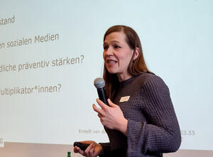 Eine Frau hält ein Mikrofon in der Hand und steht vor einer Leinwand.