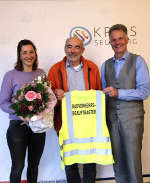 Eine Frau und zwei Männer stehen vor einer Fotowand mit der Aufschrift "Kreis Segeberg". Die Frau hält einen Blumenstrauß in den Händen, die Männer eine Warnweste mit der Aufschrift "Radverkehrsbeauftragter".