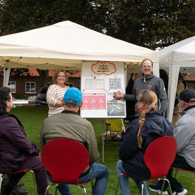 Mehrere Menschen sitzen vor einem Pavillon. Unter dem Pavillon stehen eine Frau und ein Mann mit einem Tafelschreibblock, die etwas vortragen.