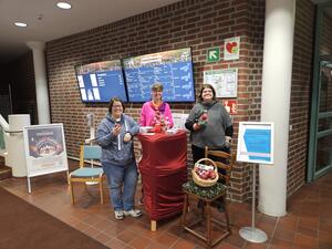 Drei Frauen stehen mit roten Äpfeln und einem Stand im Rathaus Norderstedt vor Bildschirmen.