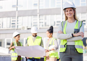 Eine junge Frau mit Helm auf dem Kopf und gelber Weste steht auf einer Baustelle.