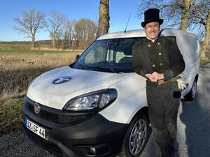 Ein Mann in schwarzer Kleidung und mit schwarzem Zylinder lehnt an einem weißen Auto und lacht freundlich in die Kamera.
