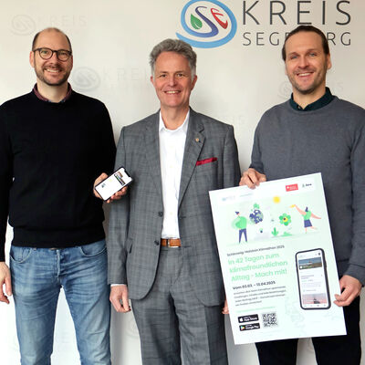Drei Männer halten ein Smartphone und ein Plakat in die Höhe.