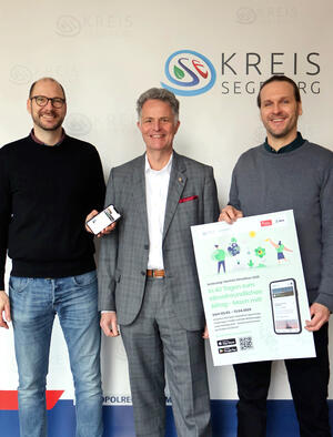 Drei Männer stehen vor einer Fotowand mit dem Logo des Kreises Segeberg und halten ein Handy sowie ein Plakat in die Kamera. Alle lächeln.