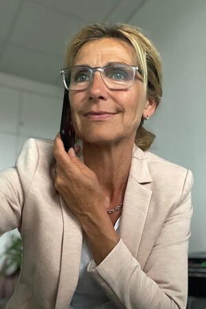 Ein blonde Frau mit Pferdeschwanz, Brille und Blazer hält einen Telefonhörer in der Hand und deutet an, auf einen Bildschirm zu schauen.