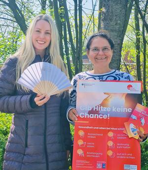 Eine blonde Frau mit langen Haaren hält einen Fächer in der Hand. Die Frau rechts neben ihr hält ein Plakat in den Händen, auf dem "Gib Hitze keine Chance!" steht.