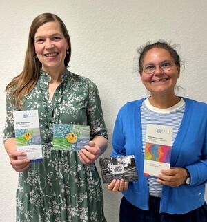 Zwei Frauen halten Flyer und Postkarten in die Kamera. Sie lächeln.