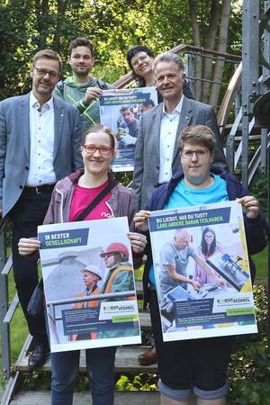Sechs Menschen lachen in die Kamera, drei von ihnen halten Plakate hoch.
